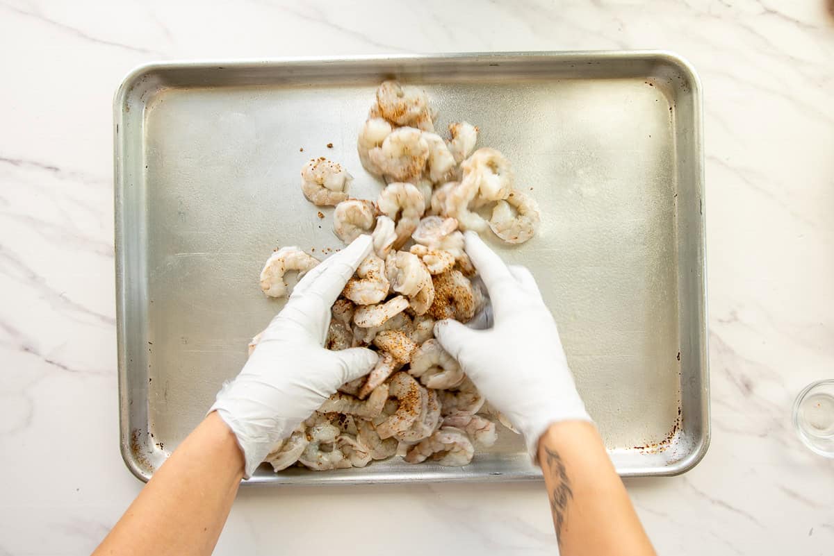 Gloved hands toss the shrimp in Tajín on a lightly oiled sheet pan.