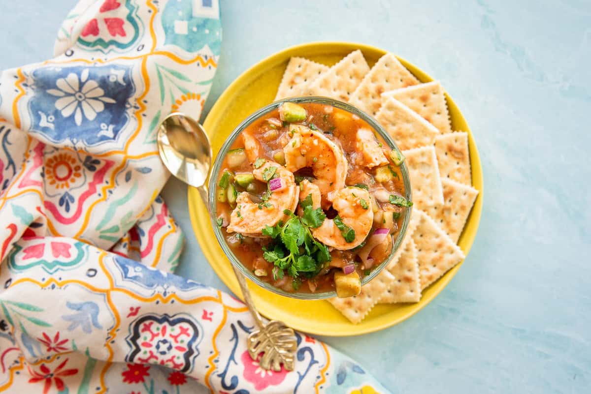 The Mexican Shrimp Cocktail is garnished with fresh cilantro and placed on a yellow plate with saltine crackers.