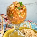 A glass goblet filled with Mexican Shrimp Cocktail on a yellow plate with saltine crackers.