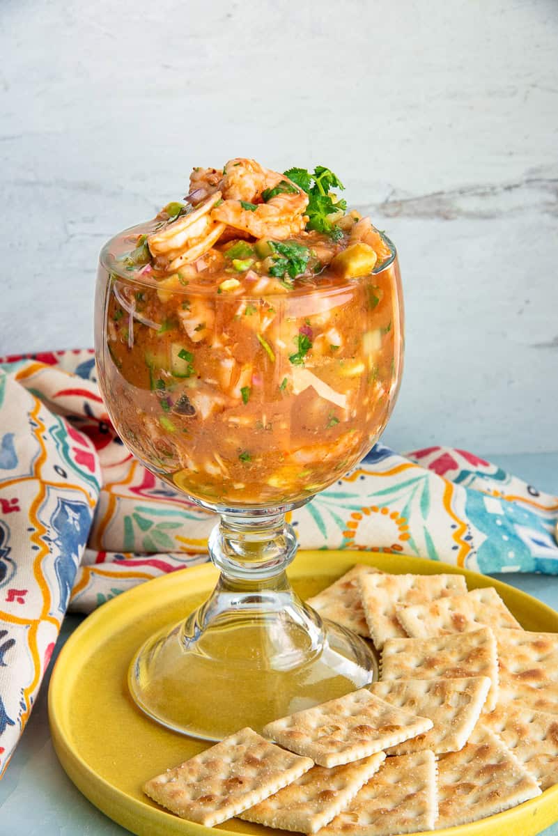 A glass goblet filled with Mexican Shrimp Cocktail on a yellow plate with saltine crackers.