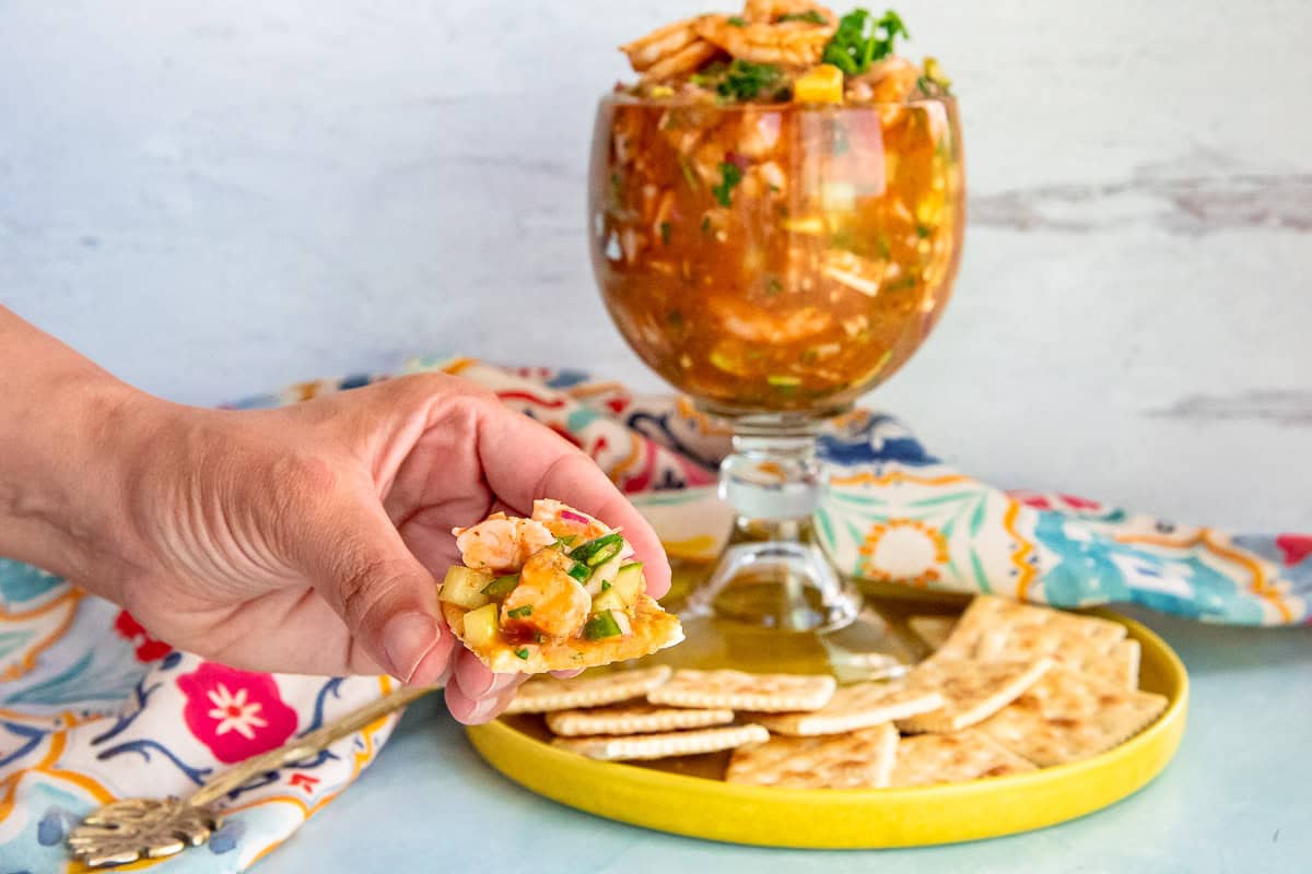 A hand holds a saltine cracker loaded with Mexican Shrimp Cocktail in front of a goblet with the rest of the recipe.
