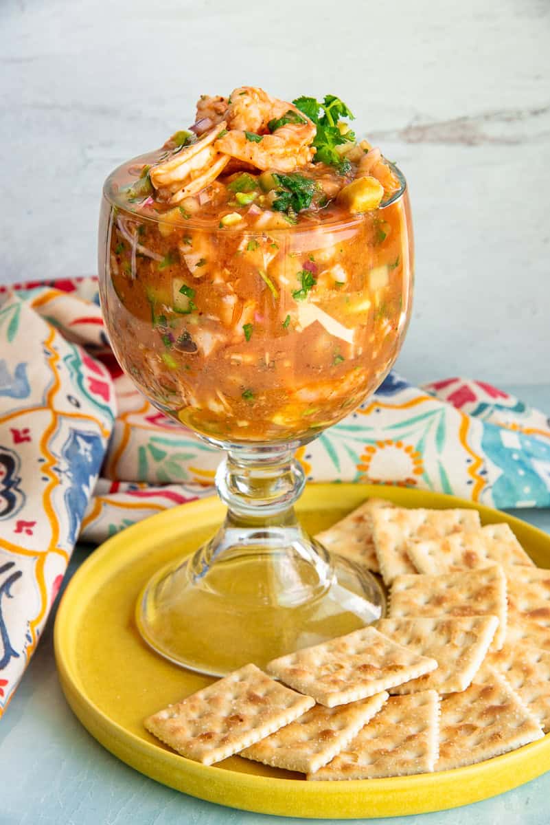 A large goblet of Mexican Shrimp Cocktail on yellow with saltine crackers in front of a multicolored kitchen towel.