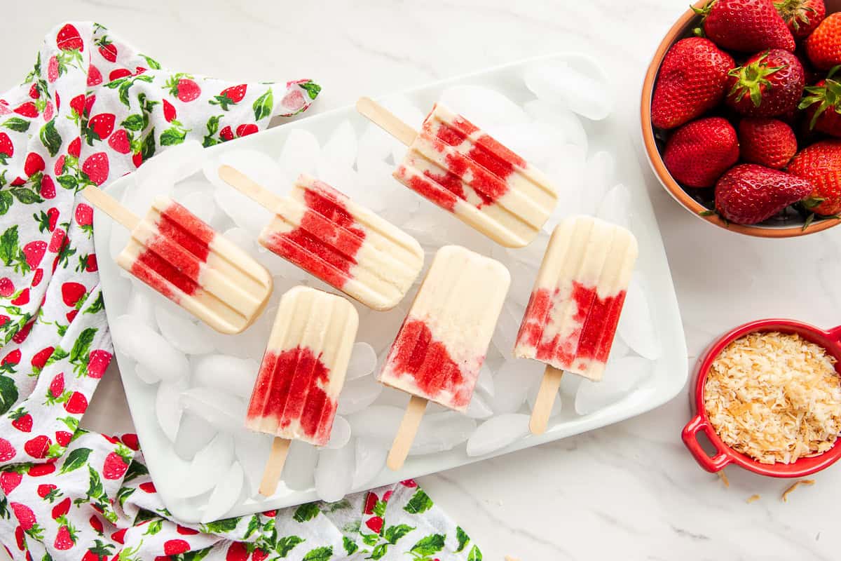 Coconut Strawberry Popsicles on a bed of ice next to a bowl of strawberries and a dish of toasted coconut flakes.