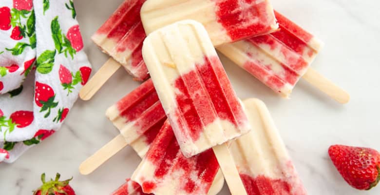 Coconut Strawberry Popsicles stacked on top of each other and surrounded by a bowl of fresh strawberries and a dish filled with toasted coconut flakes.