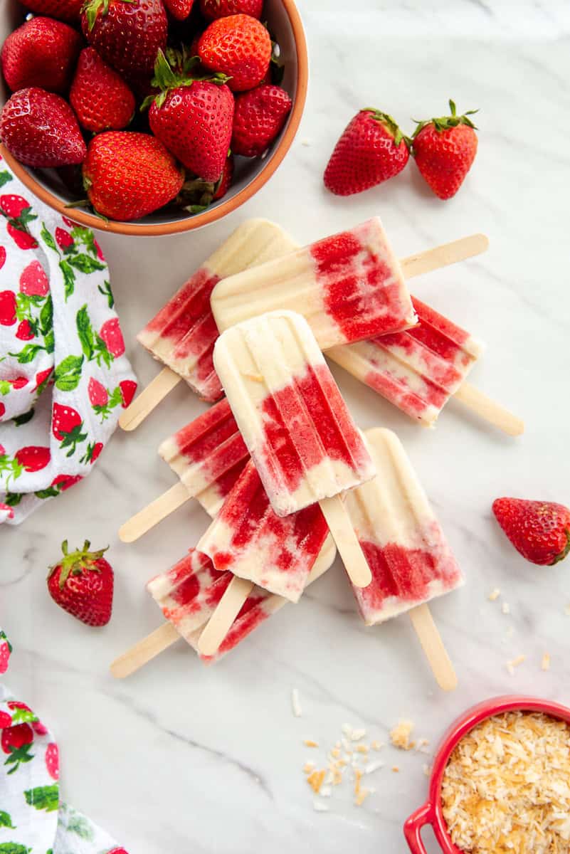 Coconut Strawberry Popsicles stacked on top of each other and surrounded by a bowl of fresh strawberries and a dish filled with toasted coconut flakes.