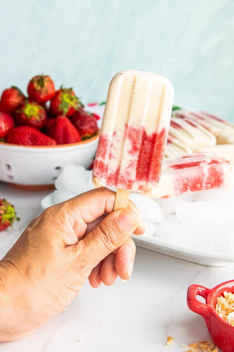 A hand holds a Coconut Strawberry Popsicle in front of a bowl of strawberries.