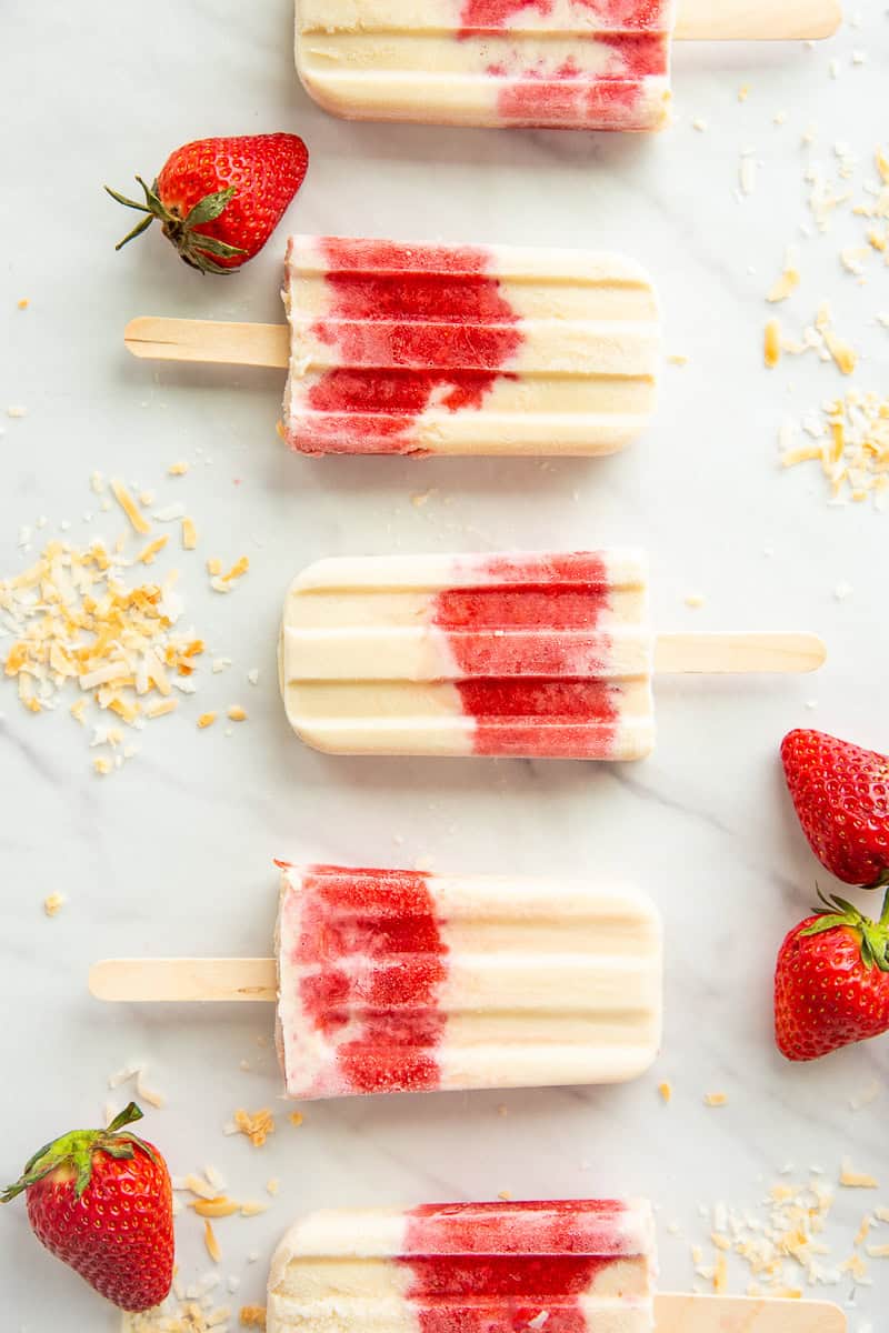 Strawberry Coconut Popsicles on a white surface lined up and surrounded by fresh strawberries and toasted coconut flakes.