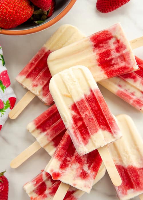 Coconut Strawberry Popsicles stacked on each other next to a kitchen towel with strawberries on it and a bowlful of strawberries.
