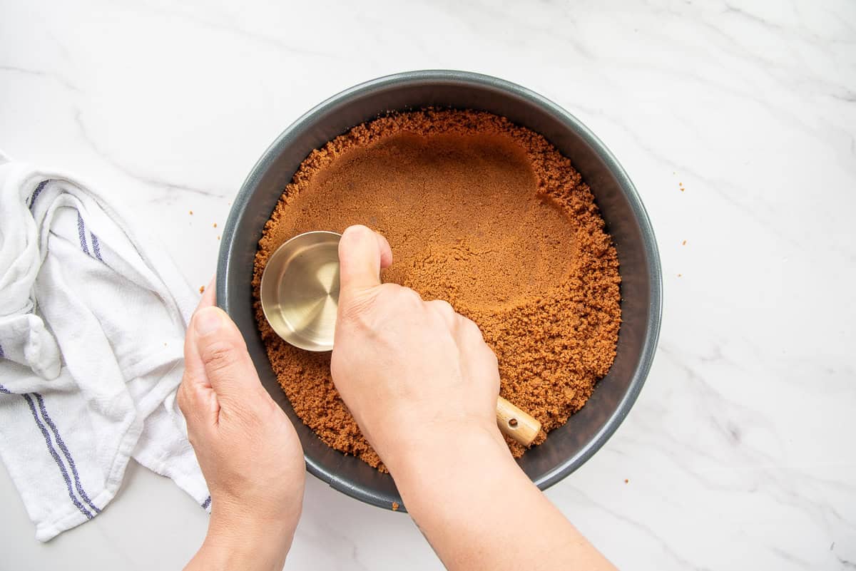 A hand uses a measuring cup to press the cookie crumbs into place in a springform pan.