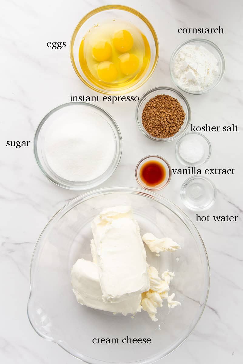 The ingredients needed to make the cheesecake are on a white countertop and labeled with black text.