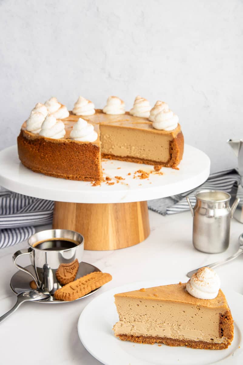 A slice of Espresso Cheesecake with Biscoff Crust on a white dessert plate next to an espresso mug and a cake stand with the rest of the cheesecake on it.