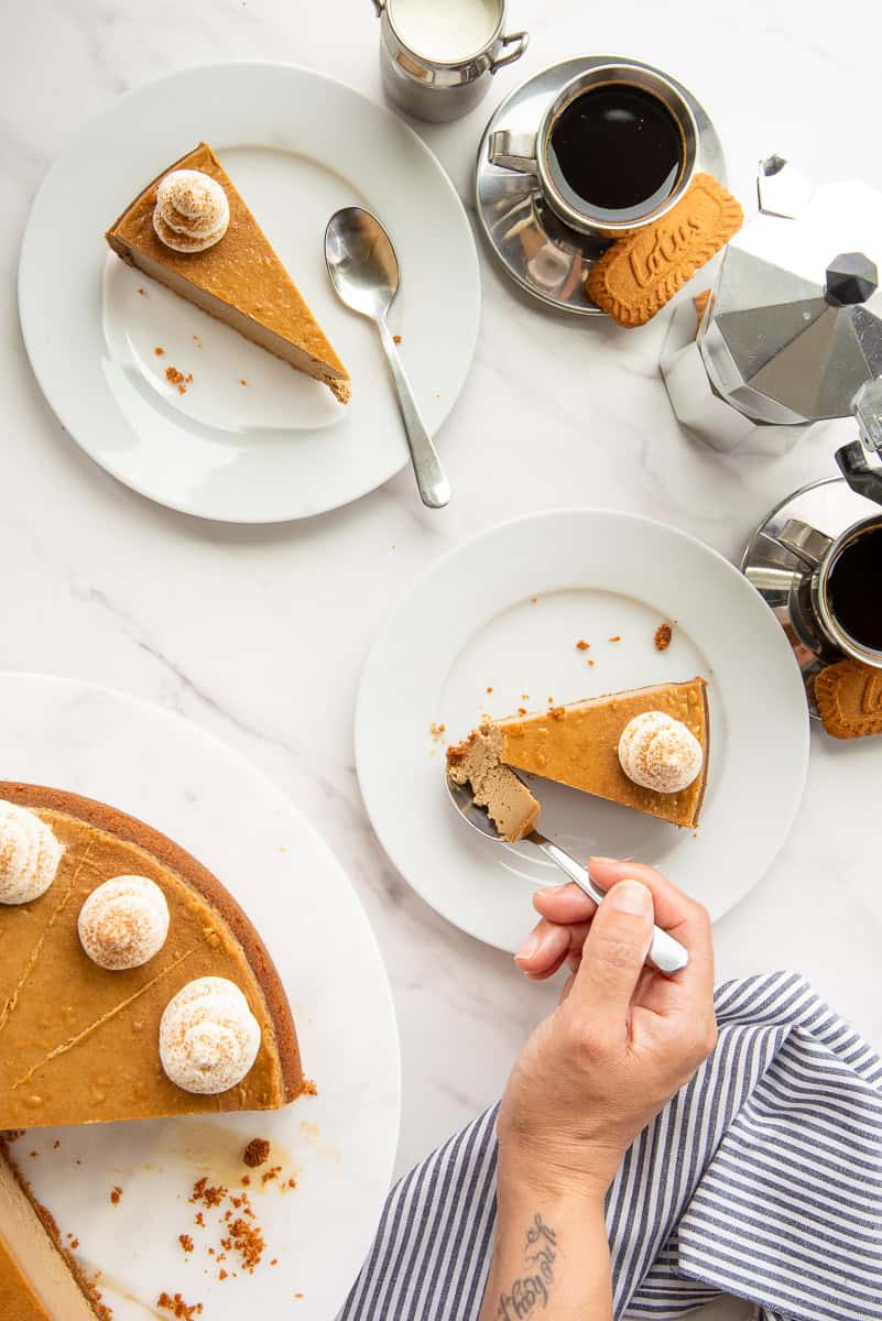 A hand removes a bite of Espresso Cheesecake with Biscoff Crust from a slice that's on a dessert plate and surrounded by cups of espresso and another plate with a slice of cheesecake on it.