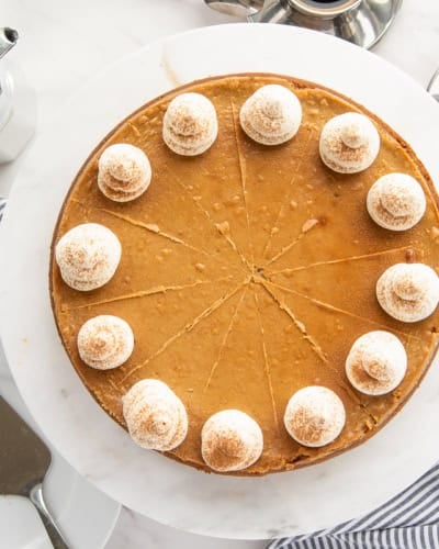 A whole Espresso Cheesecake with Biscoff Crust decorated with whipped cream next to a metal percolator and an espresso cup with a biscoff cookie next to it.