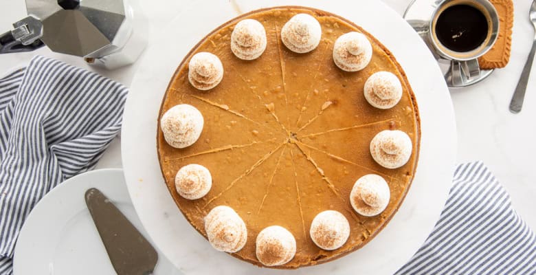 A whole Espresso Cheesecake with Biscoff Crust decorated with whipped cream next to a metal percolator and an espresso cup with a biscoff cookie next to it.