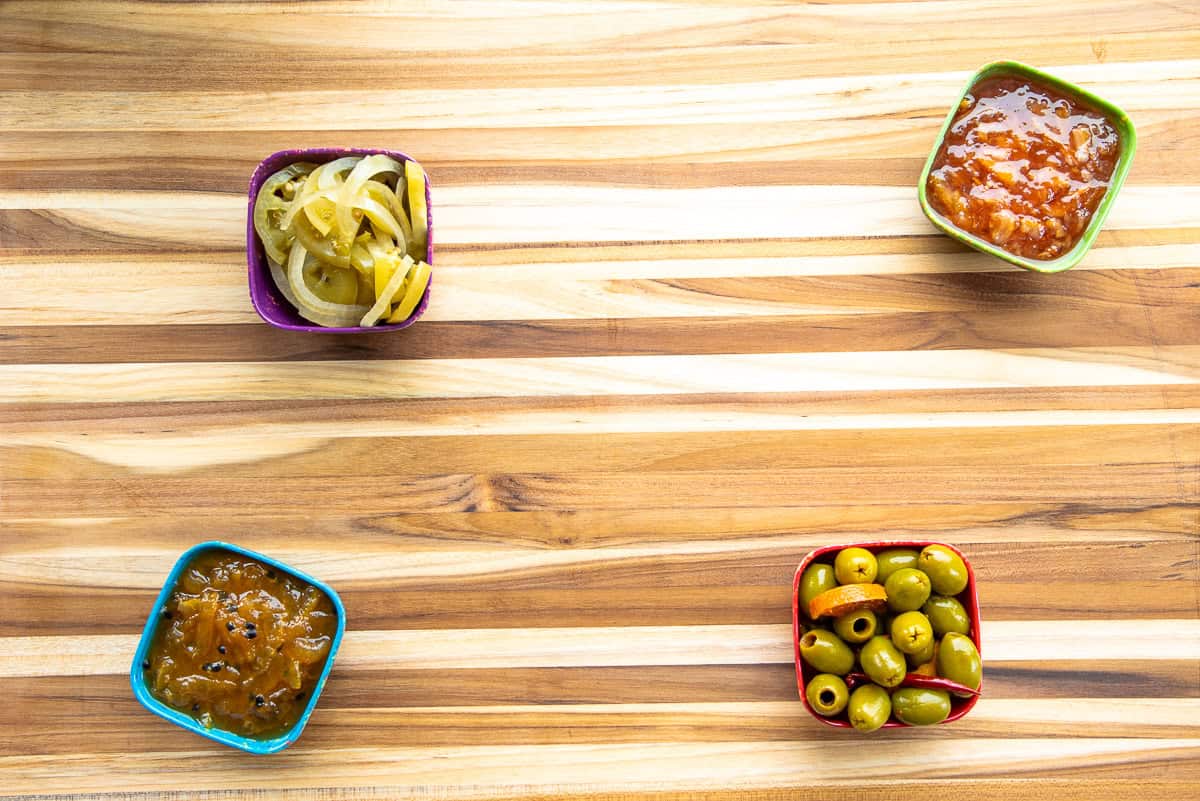 Multicolored bowls filled with condiments are placed on a wooden cutting board.