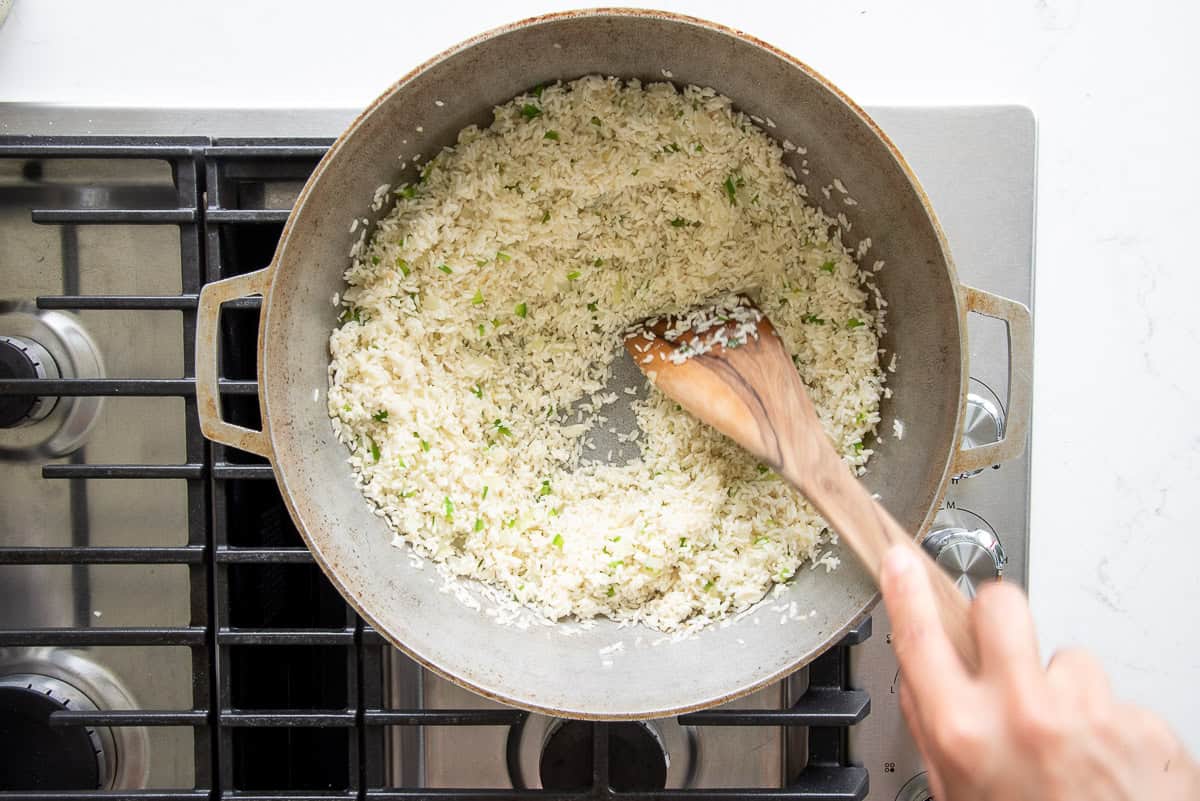 A flat wooden spoon is used to stir the rice grains into the aromatics in the pot.