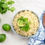 A bowl filled with Spicy Cilantro Lime Rice sits next to a wooden spoon, limes, and a bouquet of cilantro.