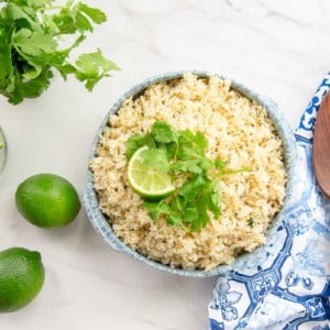 A bowl filled with Spicy Cilantro Lime Rice sits next to a wooden spoon, limes, and a bouquet of cilantro.