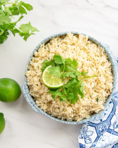 A bowl filled with Spicy Cilantro Lime Rice sits next to a wooden spoon, limes, and a bouquet of cilantro.