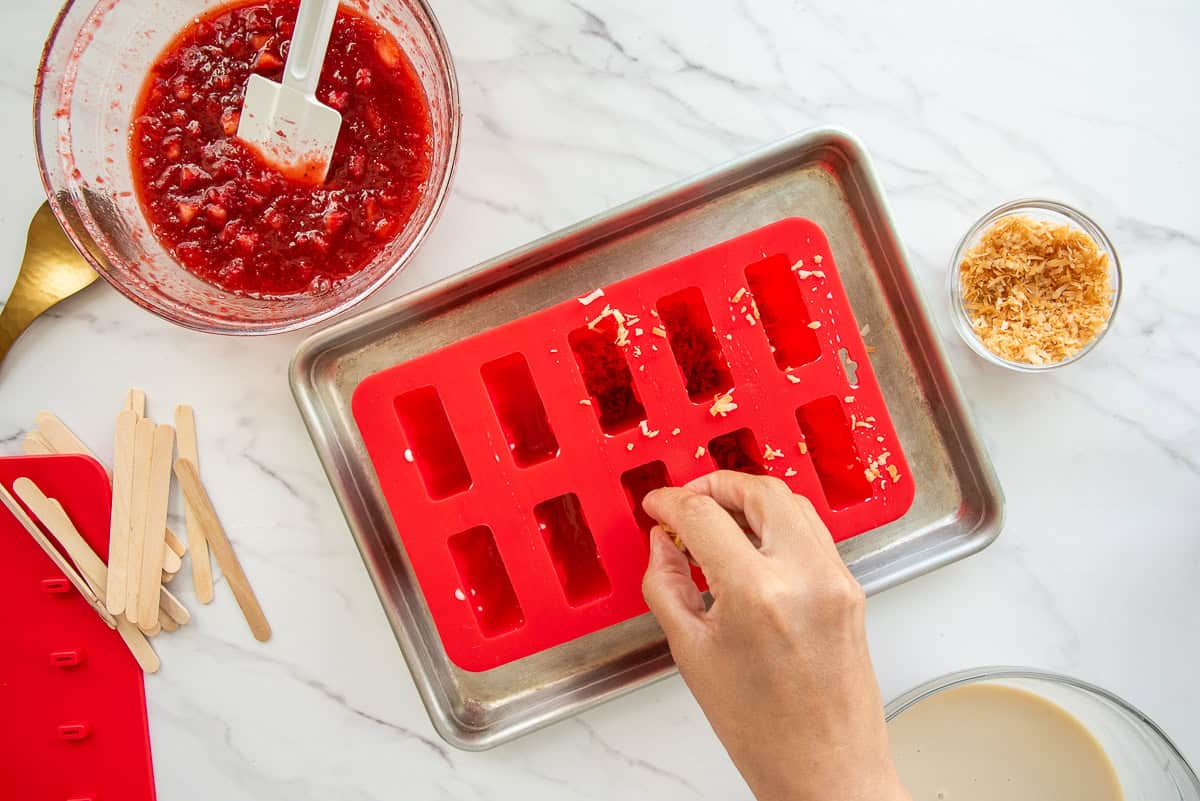 Toasted coconut flakes are sprinkled onto the semi-frozen milk mixture inside red silicone popsicle mold.