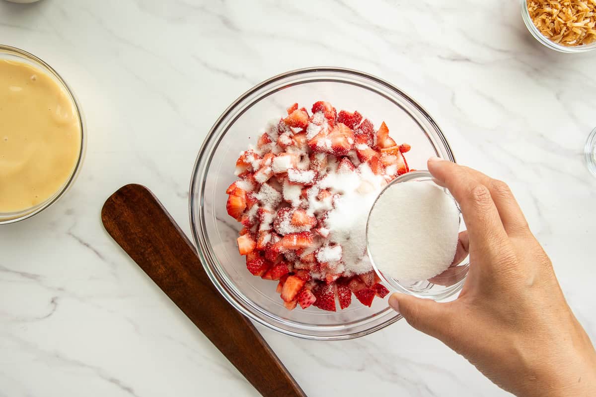 A hand pours granulated sugar from a small bowl onto the diced strawberries in another bowl.