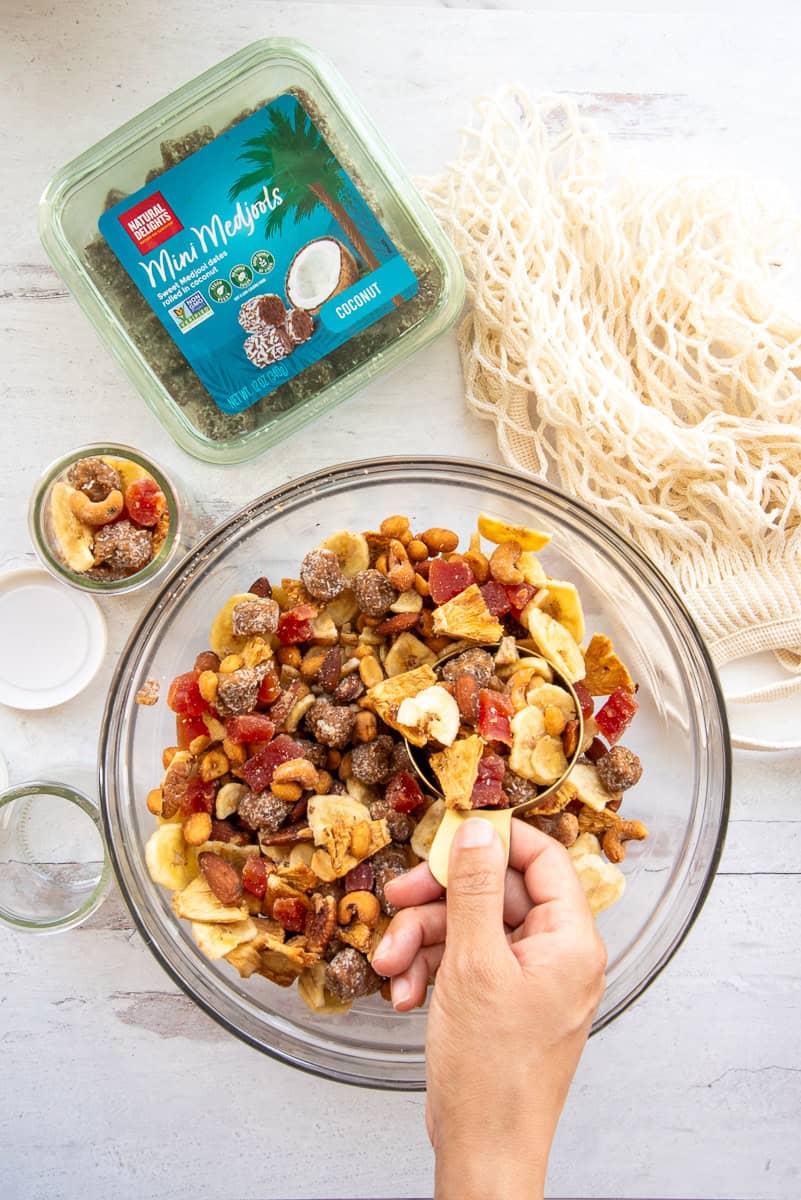 A hand is scooping up a cupful of trail mix from a glass bowl filled with it.