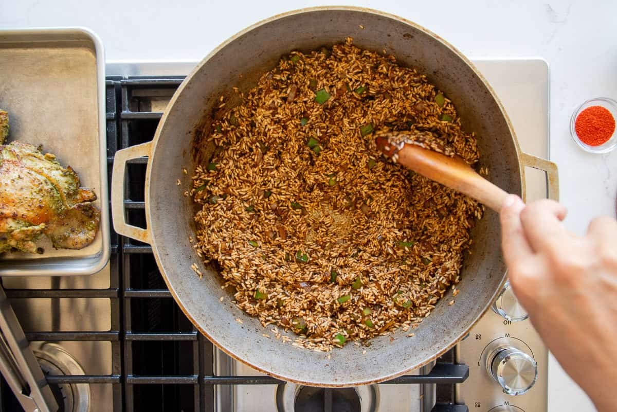 A wooden spoon is used to toss the rice in the aromatics and tomato paste mixture inside of a gray caldero.