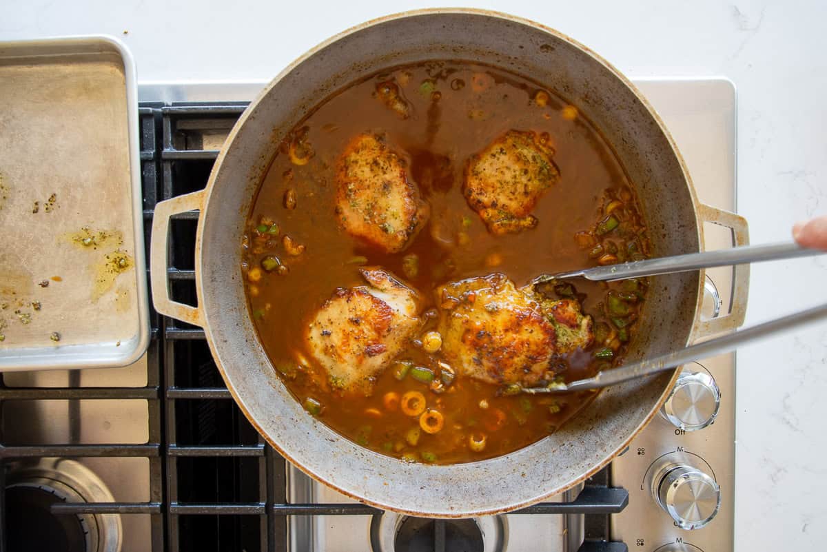 A hand uses tongs to return the seared chicken to the pot with the rice.