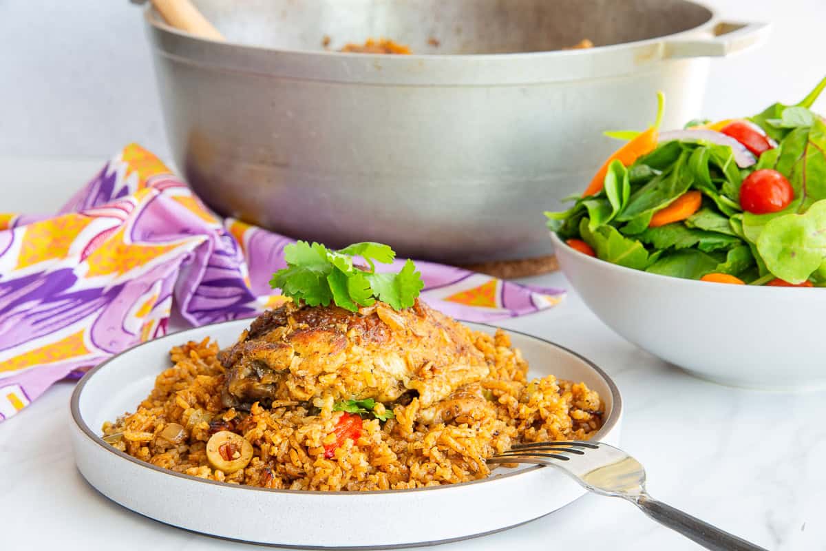 A fork is propped on a grey plate with Authentic Puerto Rican Arroz Con Pollo on it and next o a bowlful of salad and a grey caldero in the background.