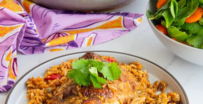 A caldero with Authentic Puerto Rican Arroz Con Pollo in it behind a grey plate with a serving of arroz con pollo and next to a white bowl filled with salad.