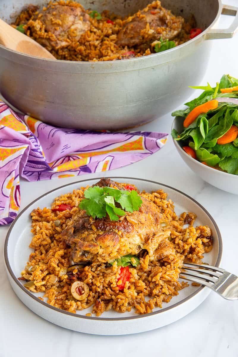 A caldero with Authentic Puerto Rican Arroz Con Pollo in it behind a grey plate with a serving of arroz con pollo and next to a white bowl filled with salad.