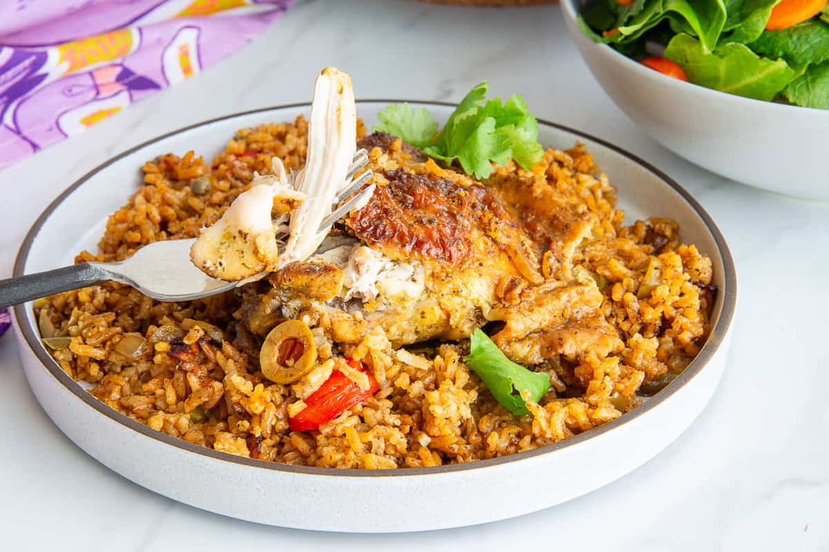 A plate of Authentic Puerto Rican Arroz Con Pollo with a fork lifting a chunk of chicken from the thigh.