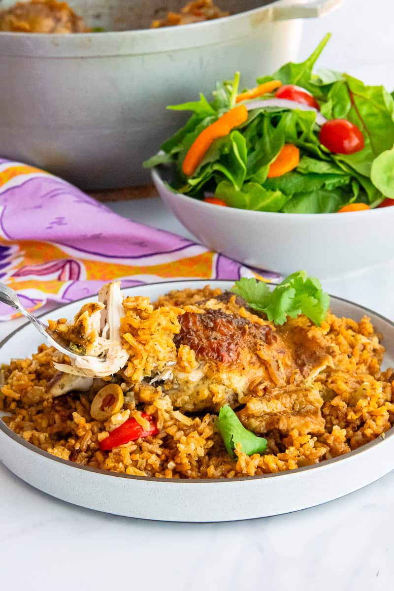 A fork lifts a chunk of chicken from a chicken thigh nestled in a bed of rice on a grey plate in front of a bowl of salad.