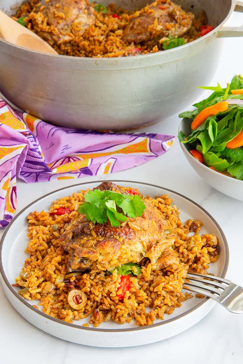 A grey plate with Authentic Puerto Rican Arroz Con Pollo on it sits next to a bowlful of salad and in front of a metal caldero.