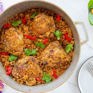 Authentic Puerto Rican Arroz Con Pollo in a caldero is garnished with roasted red pepper slices and fresh cilantro and sits next to a white bowl filled with salad and an empty grey plate.