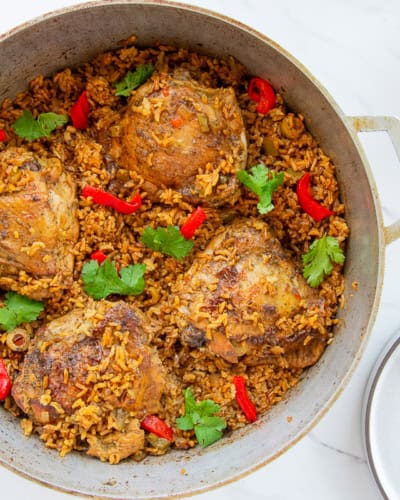 Authentic Puerto Rican Arroz Con Pollo in a caldero is garnished with roasted red pepper slices and fresh cilantro and sits next to a white bowl filled with salad and an empty grey plate.