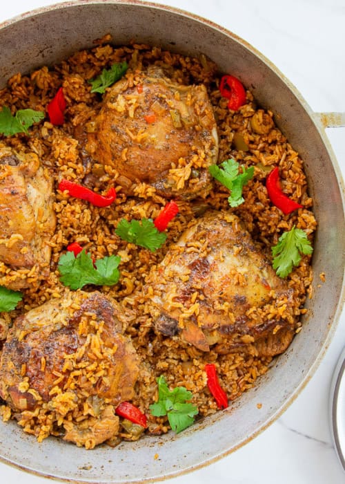 Authentic Puerto Rican Arroz Con Pollo in a caldero is garnished with roasted red pepper slices and fresh cilantro and sits next to a white bowl filled with salad and an empty grey plate.