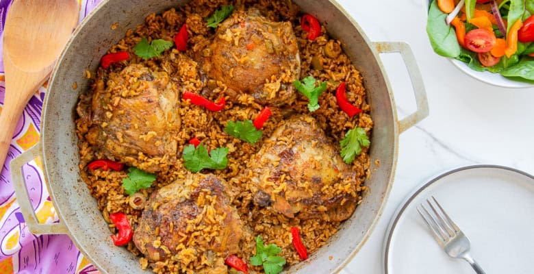 Authentic Puerto Rican Arroz Con Pollo in a caldero is garnished with roasted red pepper slices and fresh cilantro and sits next to a white bowl filled with salad and an empty grey plate.