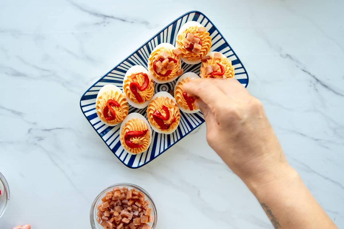 A hand sprinkles the broiled spiced ham bits onto the filled eggs.