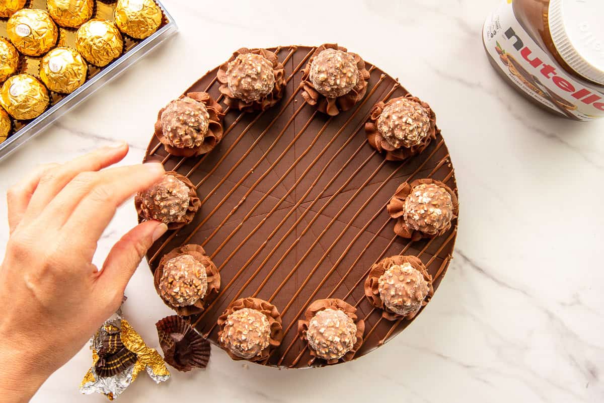 A hand places a hazelnut chocolate candy on the decorated cheesecake.