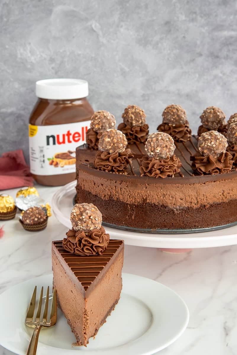 A slice of Hazelnut Chocolate Cheesecake on a plate in front of a cake stand with the rest of the cheesecake on it.