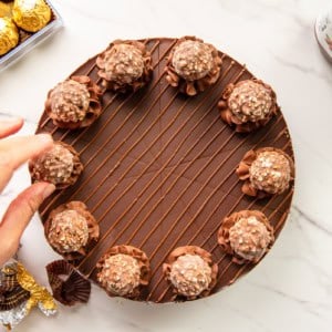 Preview of the Hazelnut Chocolate Cheesecake a hand places a candy onto a rosette piped onto the cheesecake.