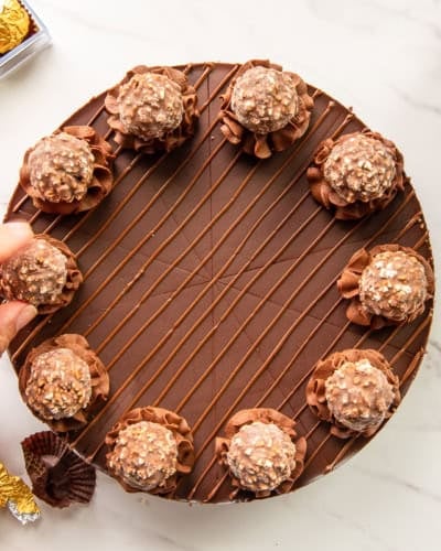 Preview of the Hazelnut Chocolate Cheesecake a hand places a candy onto a rosette piped onto the cheesecake.