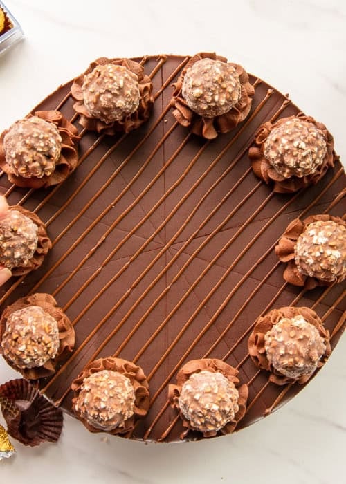 Preview of the Hazelnut Chocolate Cheesecake a hand places a candy onto a rosette piped onto the cheesecake.