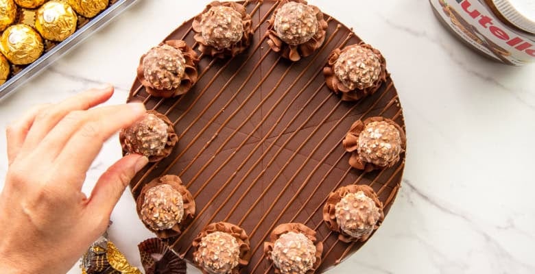 Preview of the Hazelnut Chocolate Cheesecake a hand places a candy onto a rosette piped onto the cheesecake.