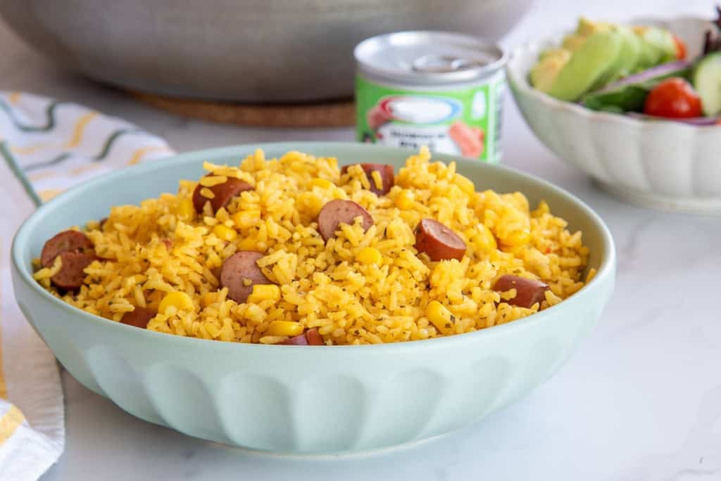 A green bowl filled with Arroz con Salchichas y Maiz in front of a tin of Vienna sausages and a bowl filled with salad.