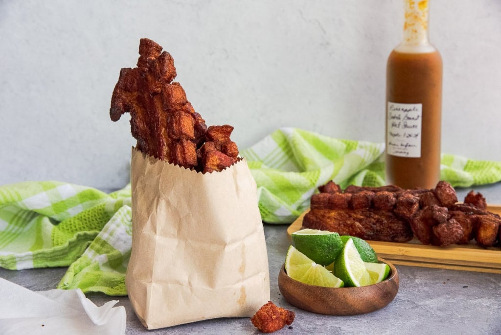 Chicharrones de Cerdo in a brown paper bag next to a bowl of lime wedges and a bottle of hot sauce.