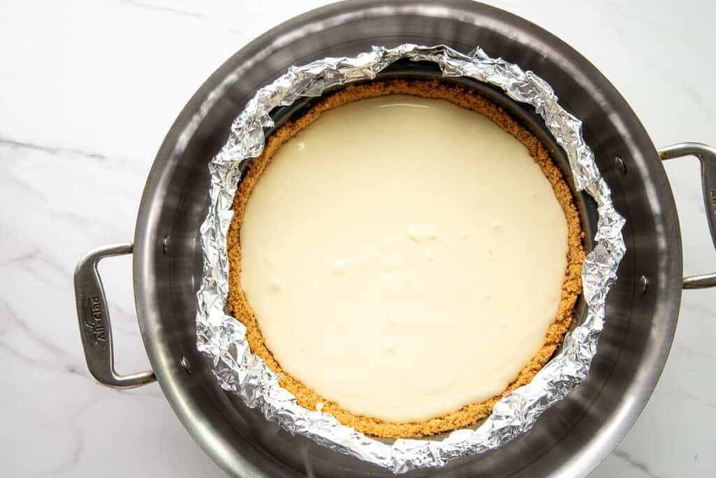 The unbaked cheesecake in a pan, wrapped in foil and set in a bain-marie inside a larger pan.
