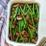 Haricot Verts with Mushrooms and Bacon is a white rectangular serving dish nect to a blue and white striped kitchen towel and a pair of serving tongs.