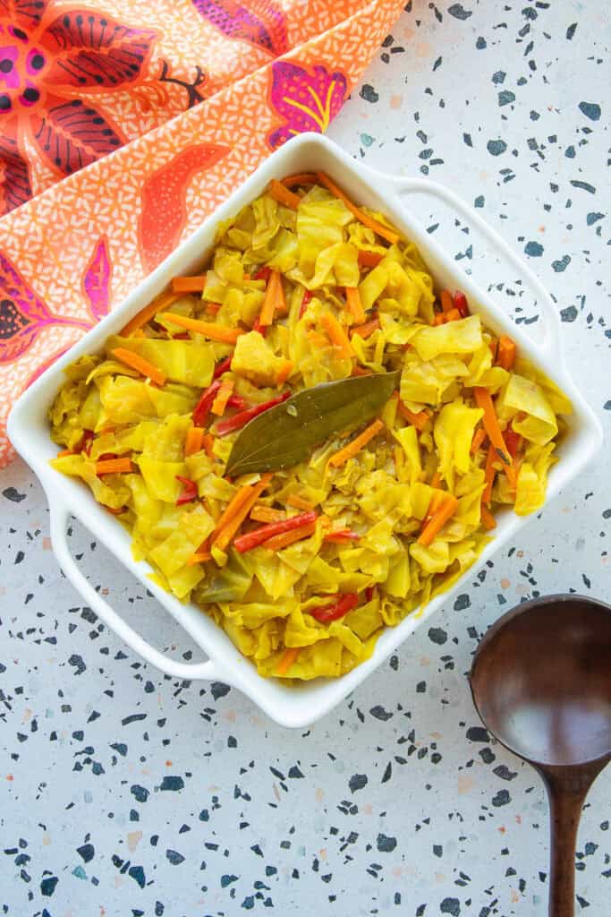 Caribbean Steamed Cabbage served in a white ceramic dish next to an orange kitchen towel and a dark wooden spoon.